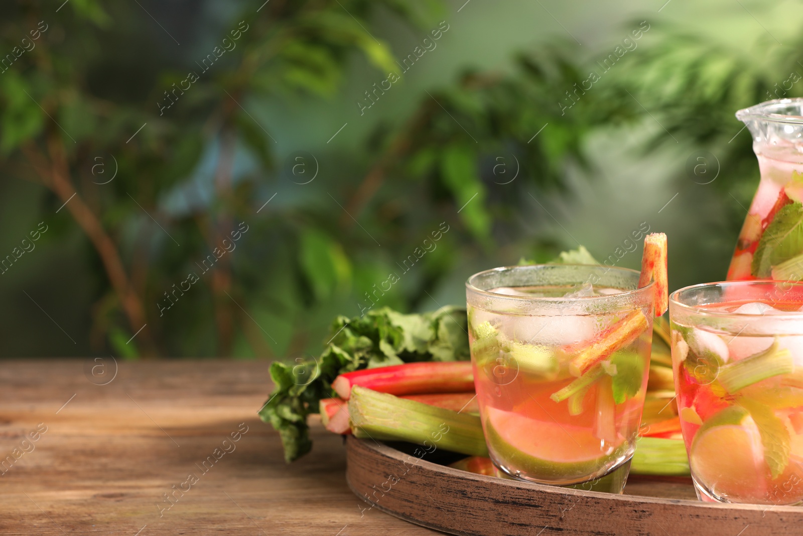 Photo of Glasses and jug of tasty rhubarb cocktail with citrus fruits on wooden table outdoors, space for text