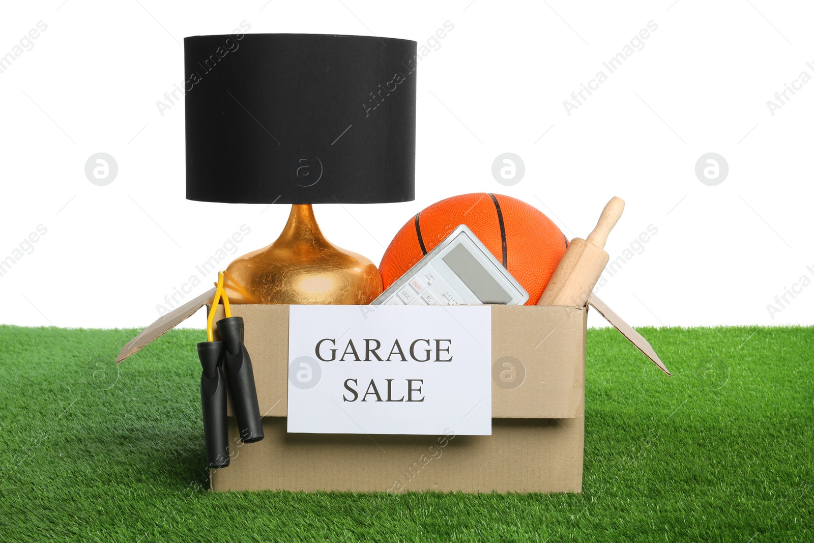 Photo of Different stuff in box with sign Garage Sale on green grass against white background