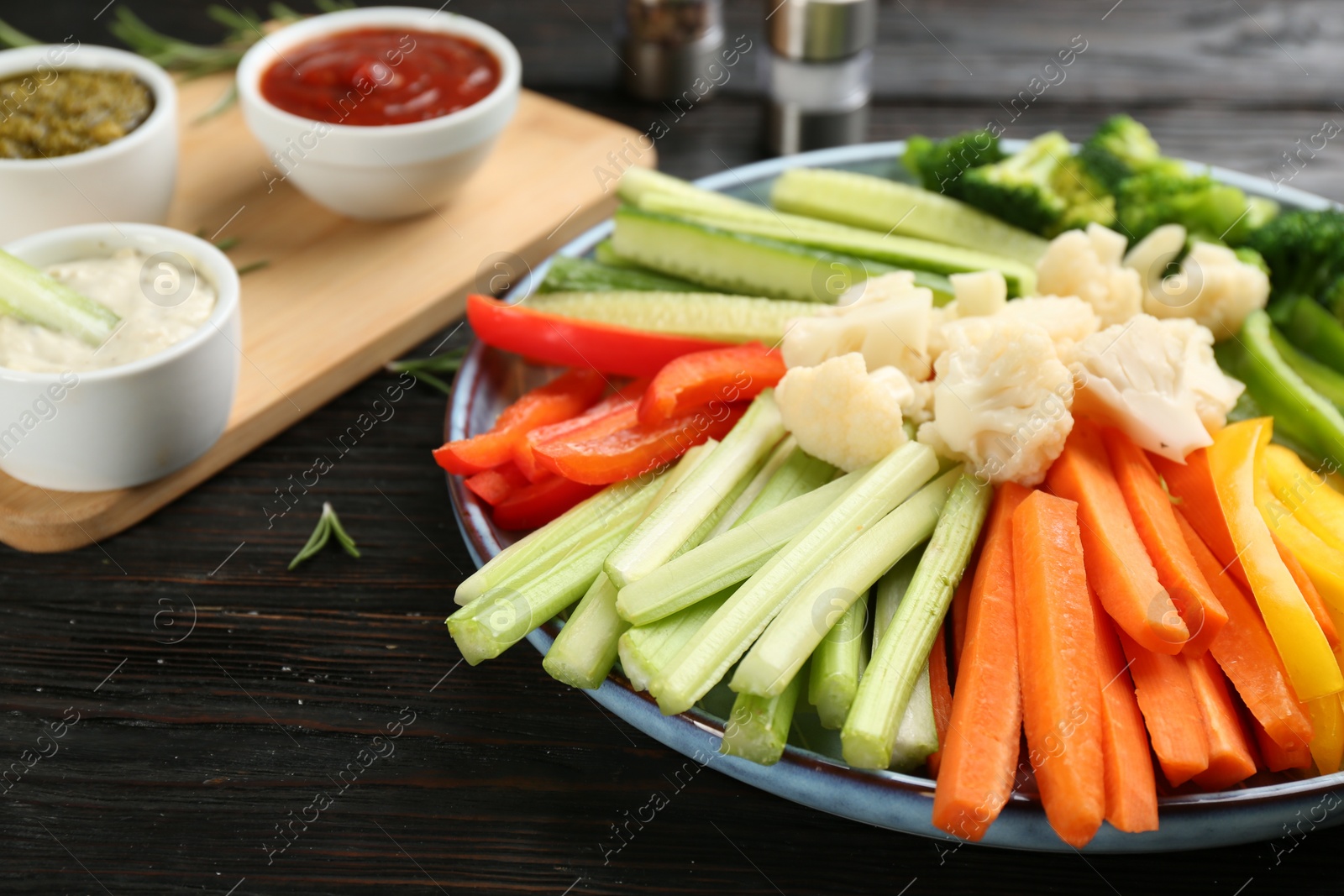 Photo of Plate with celery sticks, other vegetables and different dip sauces on dark wooden table, closeup