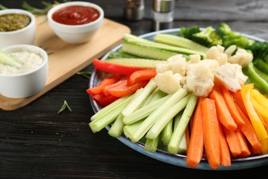 Photo of Plate with celery sticks, other vegetables and different dip sauces on dark wooden table, closeup