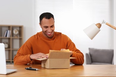 Happy young man opening parcel at table indoors. Internet shopping