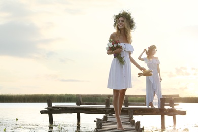 Photo of Young women wearing wreaths made of beautiful flowers on pier near river