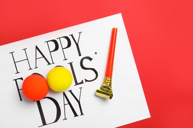 Photo of Sheet of paper with phrase Happy Fools' Day, clown noses and party blower on red background, flat lay