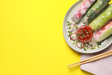 Delicious spring rolls served with sauce on yellow background, flat lay. Space for text