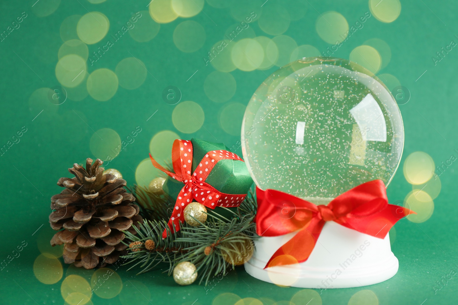 Image of Beautiful Christmas snow globe with red bow, fir cone and branch on green background, bokeh effect