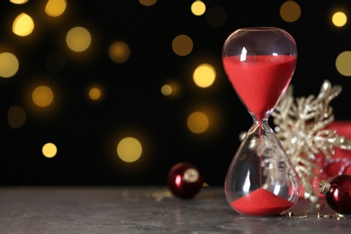 Photo of Hourglass with flowing sand and decor on grey table against blurred lights, space for text. Christmas countdown