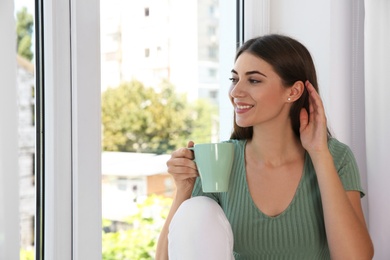 Photo of Beautiful young woman with cup of drink on window sill indoors