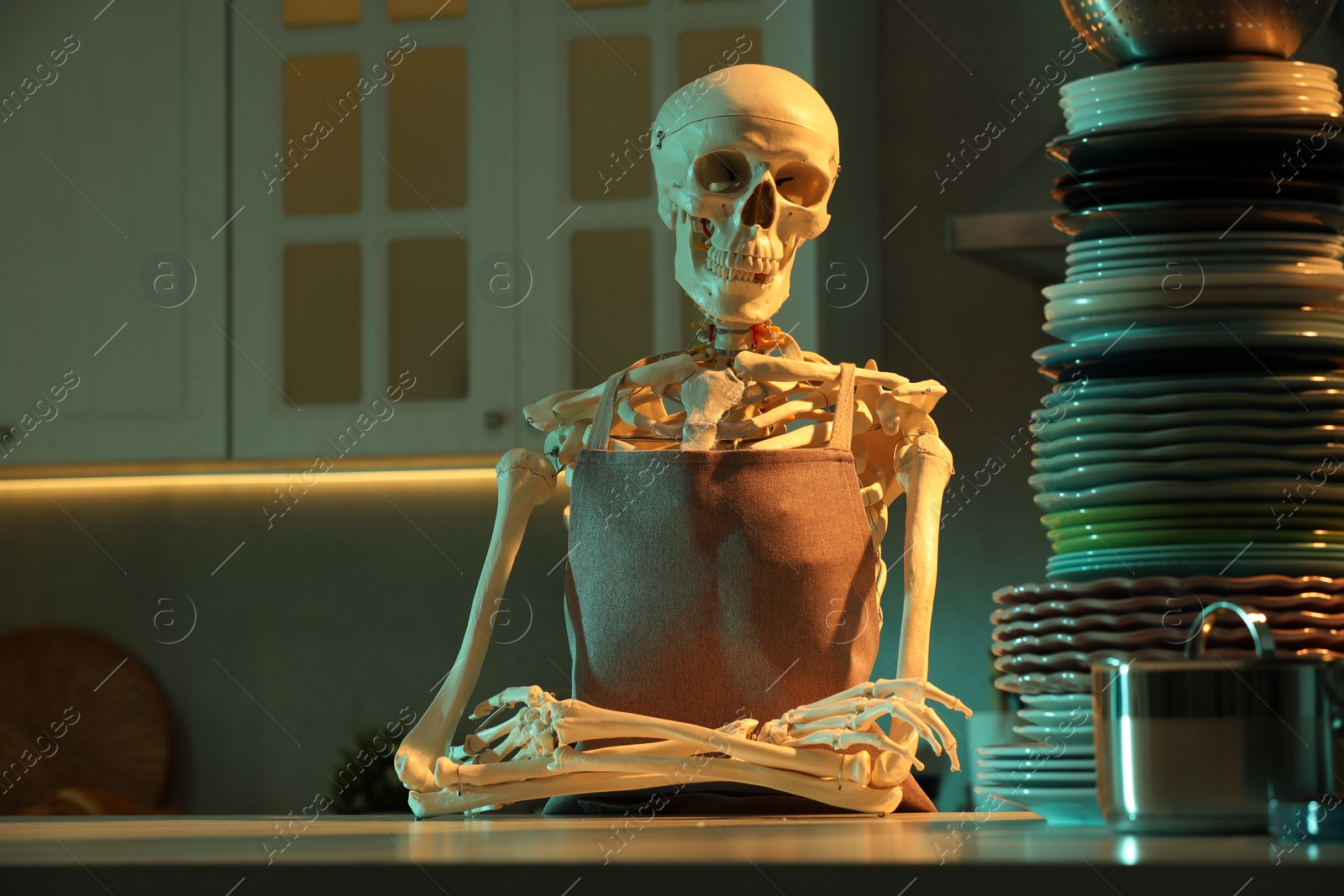 Photo of Human skeleton sitting at white marble table with clean dishware in kitchen at night