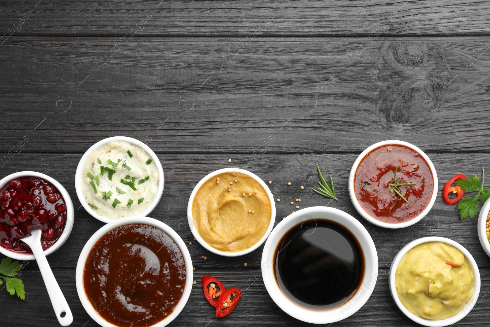 Photo of Different tasty sauces in bowls, parsley, chili pepper and rosemary on black wooden table, flat lay. Space for text