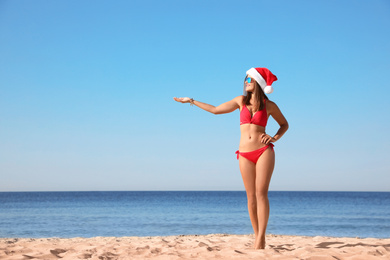 Young woman wearing Santa hat and bikini on beach, space for text. Christmas vacation