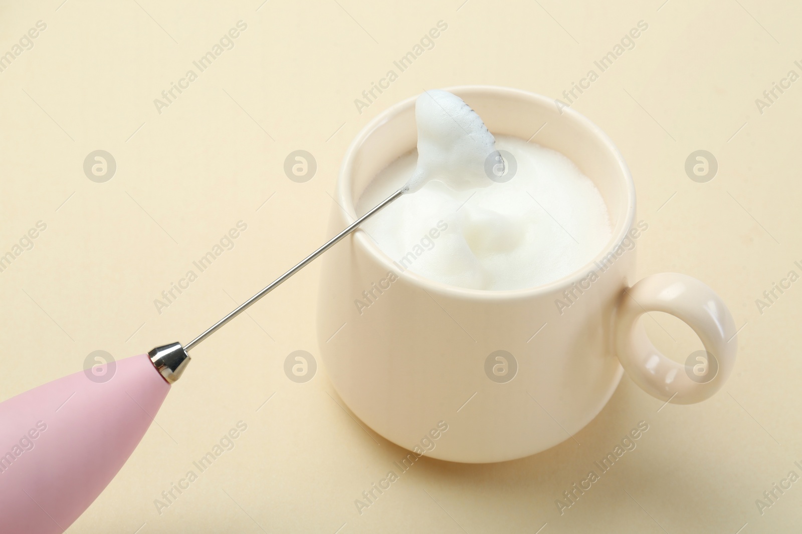 Photo of Mini mixer (milk frother) and cup of whipped milk on beige background, closeup