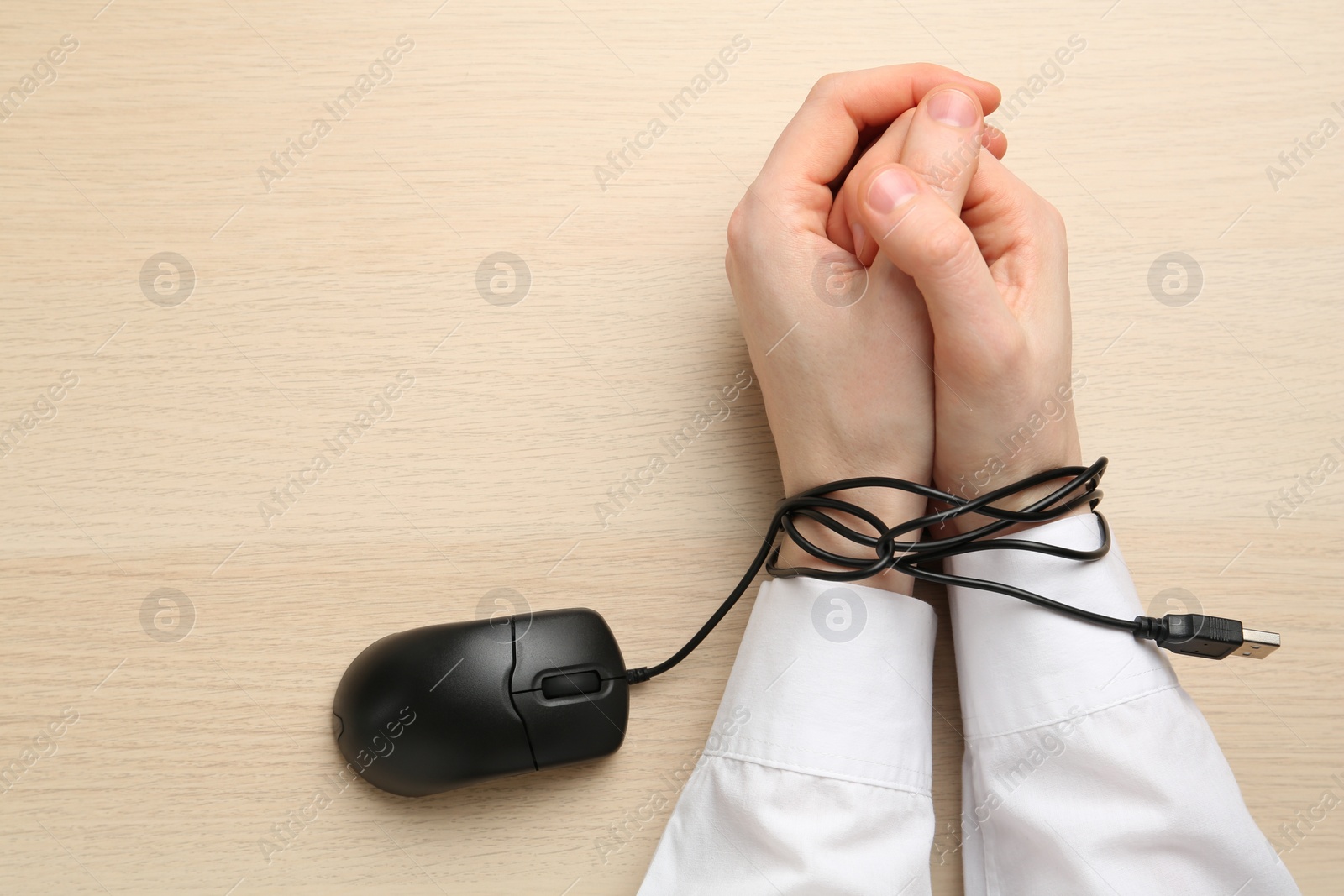 Photo of Man showing hands tied with computer mouse cable at wooden table, top view. Internet addiction