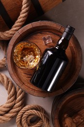 Whiskey with ice cubes in glass, bottle, wooden barrels and rope on grey marble table, flat lay