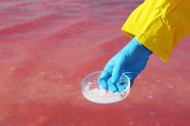 Photo of Laboratory worker with Petri dish taking sample from pink lake for analysis, closeup