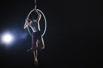 Photo of Young woman performing acrobatic element on aerial ring against dark background. Space for text