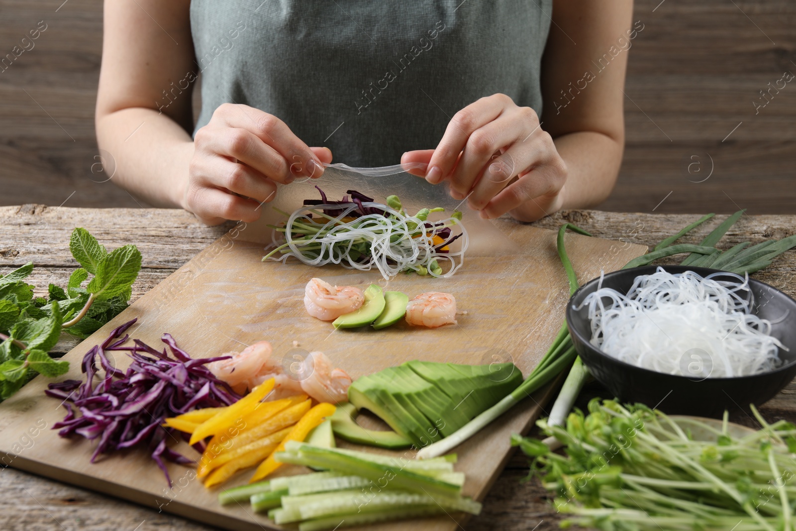 Photo of Woman wrapping spring roll at wooden table with products, closeup