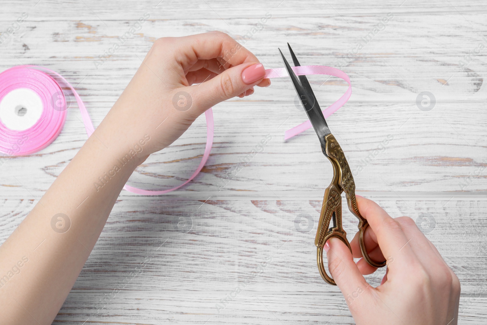 Photo of Woman cutting pink ribbon with scissors at white wooden table, top view