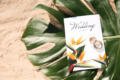 Photo of Wedding invitation, gold rings and palm leaf on sandy beach, flat lay