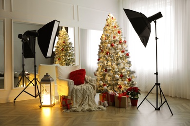 Photo of Cozy festive photo zone with armchair, Christmas tree and professional equipment indoors