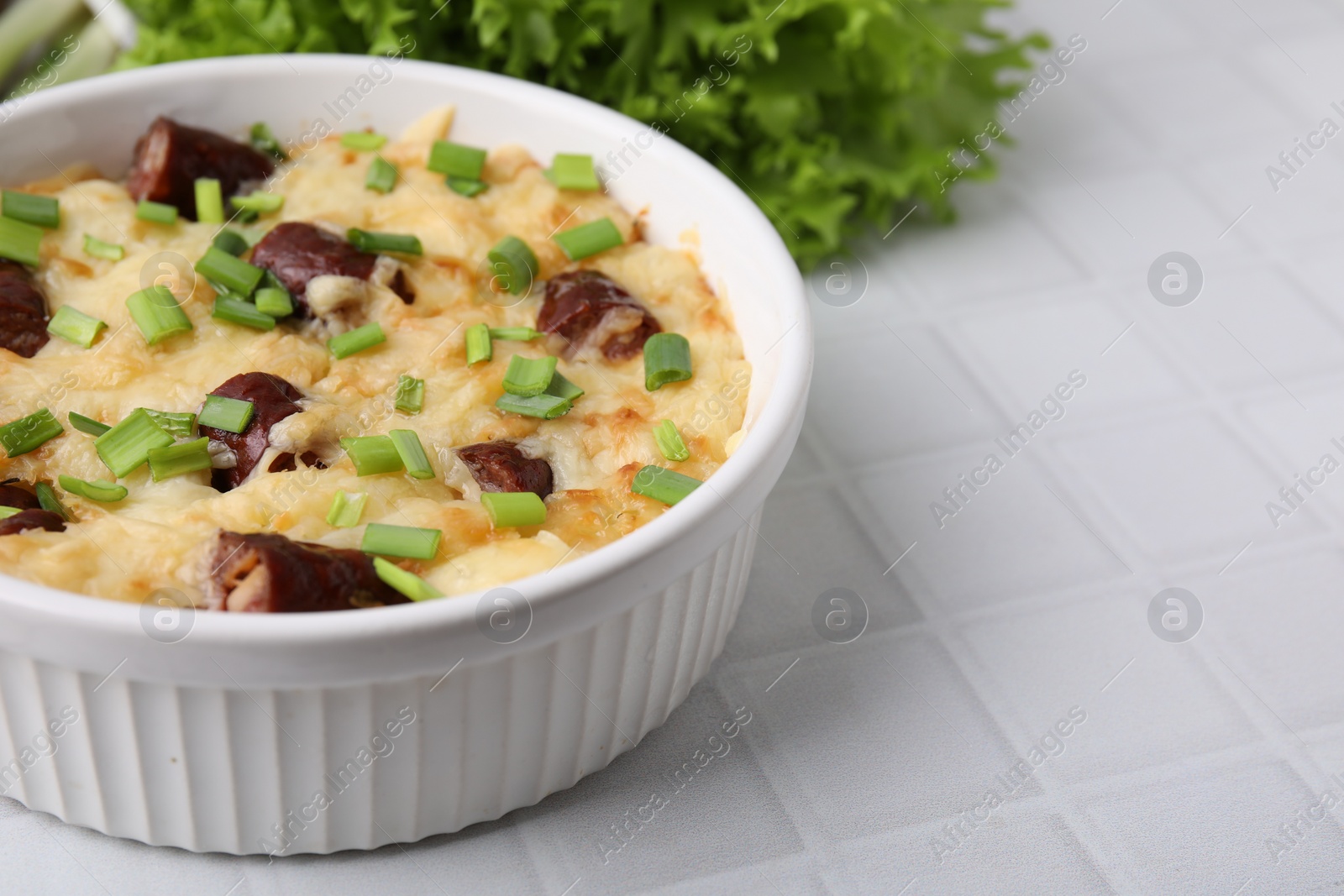 Photo of Tasty sausage casserole with green onion in baking dish on white tiled table, closeup. Space for text