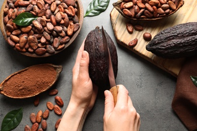 Woman cutting ripe cocoa pod over grey table with products, top view