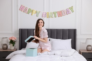Beautiful young woman with headband opening gift box on bed in room. Happy Birthday