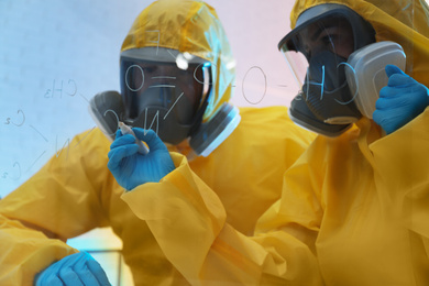 Scientists in chemical protective suits writing formula on glass board at laboratory. Virus research