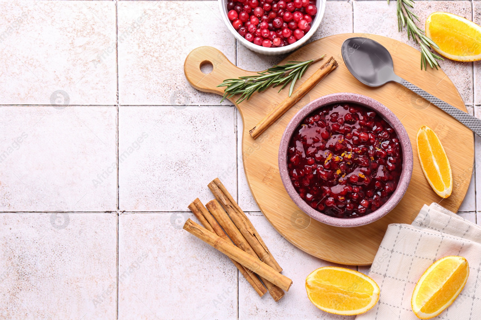 Photo of Tasty cranberry sauce in bowl and ingredients on white tiled table, flat lay. Space for text