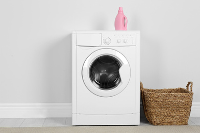 Modern washing machine with detergent and laundry basket near white wall