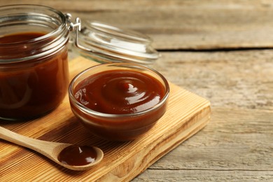 Photo of Tasty barbeque sauce in bowl, jar and spoon on wooden table, closeup. Space for text