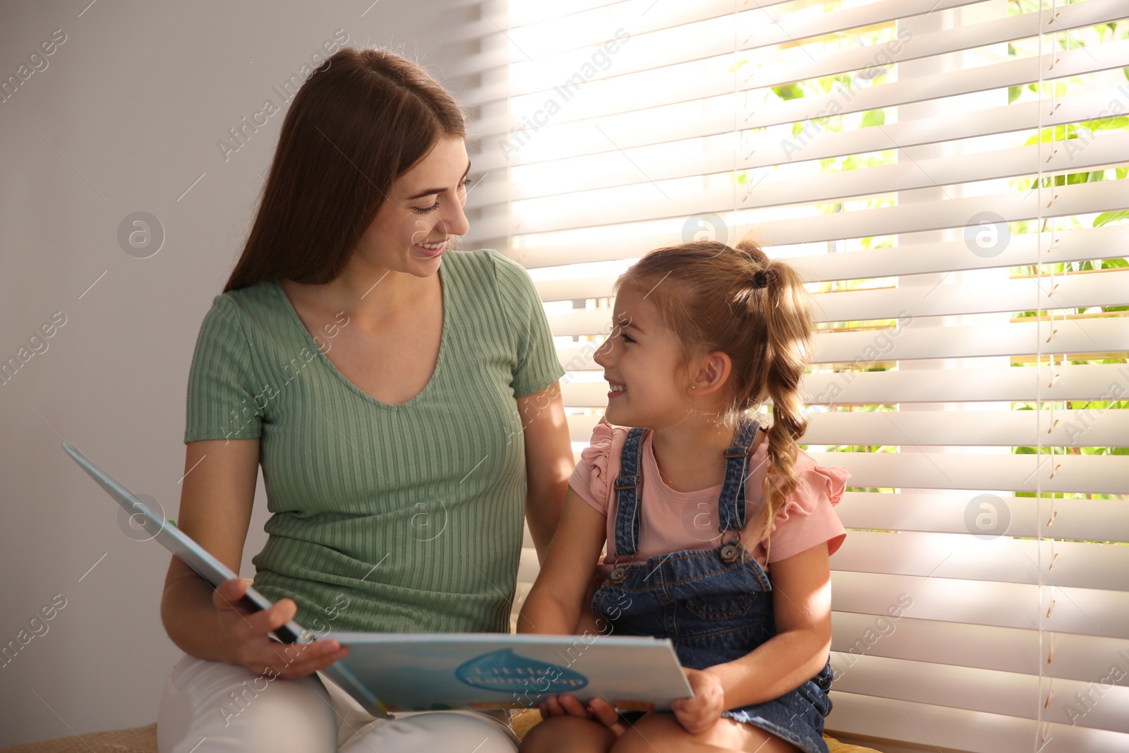 Photo of Young woman and her daughter reading book near window at home