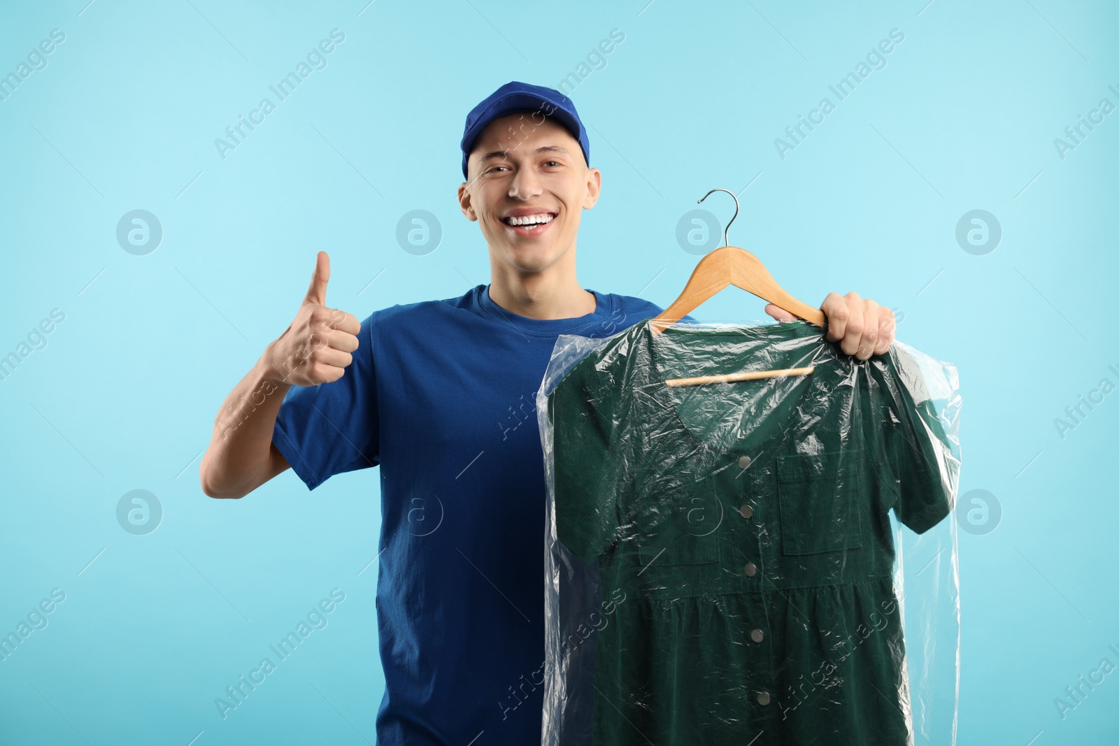 Photo of Dry-cleaning delivery. Happy courier holding dress in plastic bag and showing thumbs up on light blue background