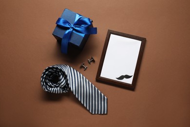 Photo of Empty photo frame, gift box and men accessories on brown background, flat lay with space for text. Father's day celebration