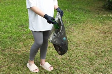 Woman with trash bag picking up glass bottle outdoors, closeup. Recycling concept
