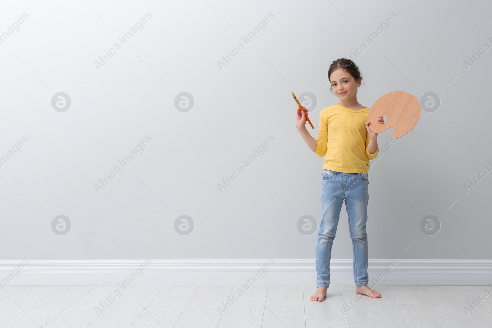 Photo of Little girl with brush and paint palette near light grey wall indoors. Space for text