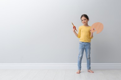 Photo of Little girl with brush and paint palette near light grey wall indoors. Space for text