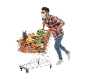 Young man in medical mask with shopping cart full of groceries on white background