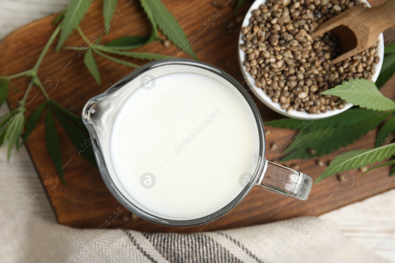 Photo of Glass jug with fresh hemp milk, seeds and leaves on wooden board, flat lay