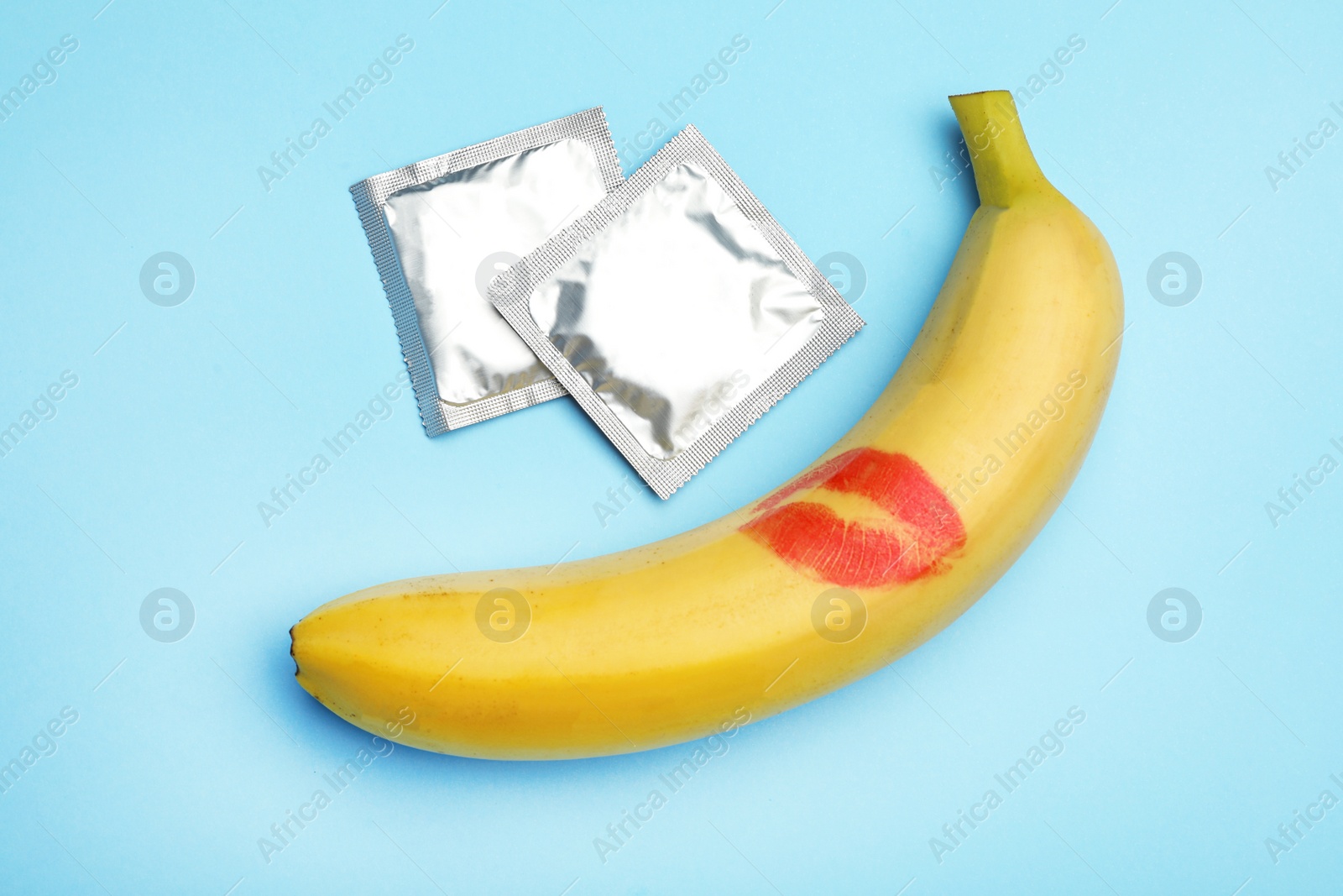 Photo of Condoms and banana with lipstick kiss mark on light blue background, flat lay. Safe sex