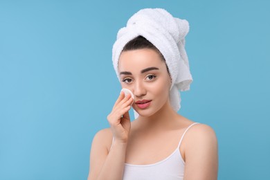 Photo of Beautiful woman in terry towel removing makeup with cotton pad on light blue background, space for text