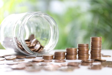 Overturned glass jar and metal coins on white table against blurred green background