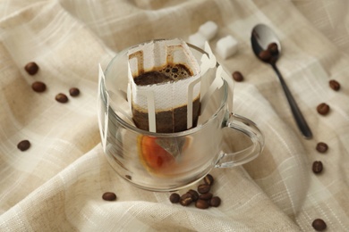 Photo of Glass cup with drip coffee bag and beans on beige fabric, closeup