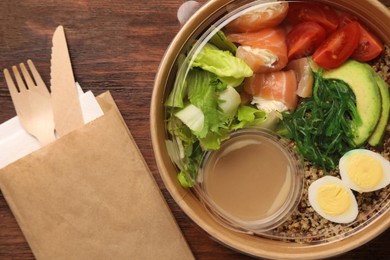 Tasty food in container with knife and fork on wooden table, flat lay