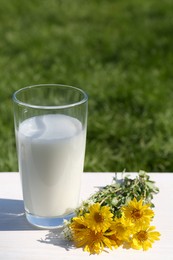 Photo of Glass of fresh milk and flowers on white wooden table outdoors