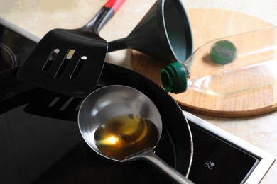 Photo of Frying pan of used cooking oil with ladle, empty bottle and funnel in kitchen, closeup