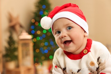 Cute little baby in Christmas sweater and Santa hat at home, space for text. Winter holiday