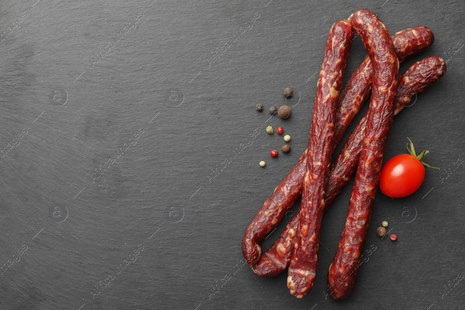 Photo of Many thin dry smoked sausages, peppercorns and tomato on black table, flat lay. Space for text
