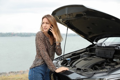 Troubled young woman talking on phone near broken car outdoors