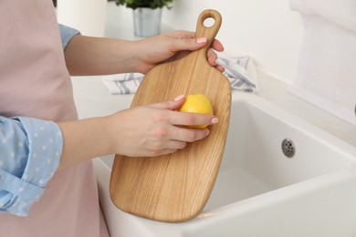 Photo of Woman rubbing wooden cutting board with lemon at sink in kitchen, closeup