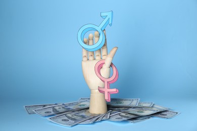 Photo of Gender pay gap. Wooden mannequin hand with symbols and dollar banknotes on light blue background